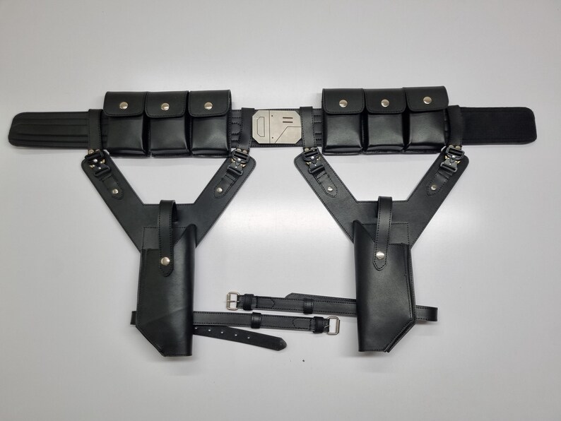 May include: A black utility belt featuring six rectangular pouches with silver snap closures, a central metallic buckle, and two integrated holsters suspended from Y-shaped straps. The assembly includes adjustable leg straps with silver buckles.