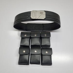 May include: A black utility belt featuring a metallic silver buckle with geometric detailing. The belt is accompanied by six detachable black rectangular pouches, each secured with a single silver-toned snap button closure on the front flap.