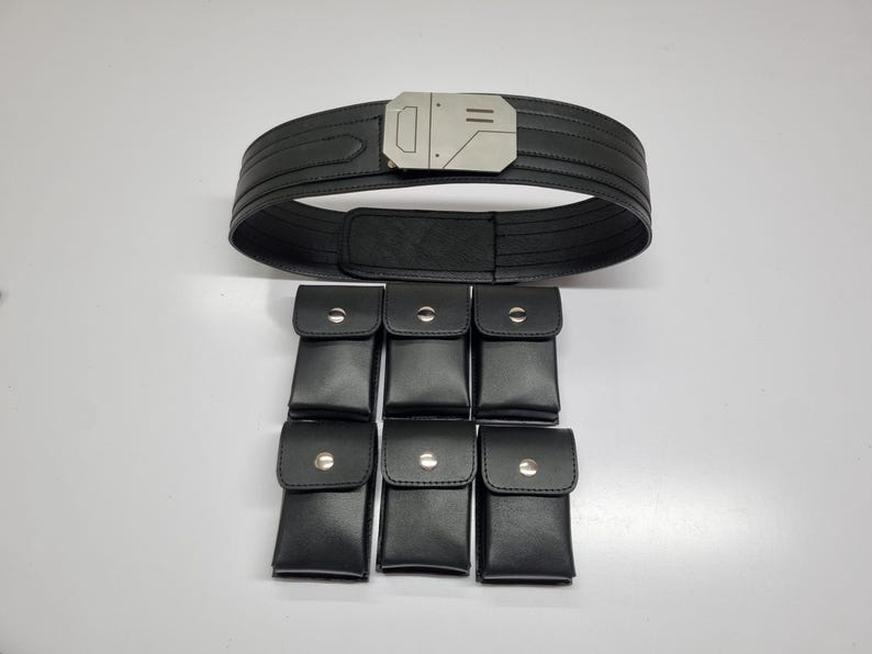 May include: A black utility belt featuring a metallic silver buckle with geometric detailing. The belt is accompanied by six detachable black rectangular pouches, each secured with a single silver-toned snap button closure on the front flap.