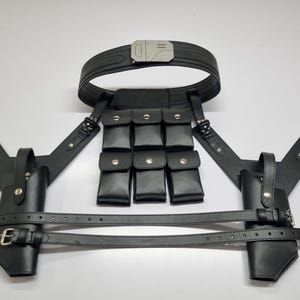 May include: A black tactical harness system featuring a wide belt with a metallic buckle, two holsters with adjustable straps, and a central panel containing six small rectangular pouches with silver-toned snap closures, all constructed from black material.