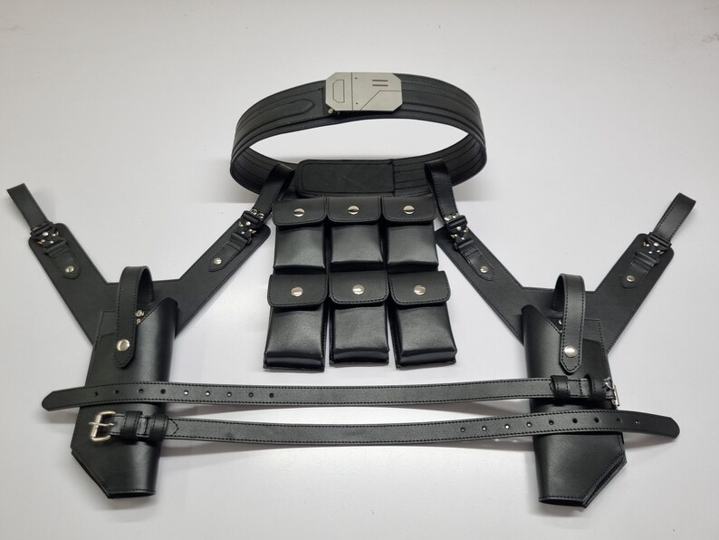 May include: A black tactical harness system featuring a wide belt with a metallic buckle, two holsters with adjustable straps, and a central panel containing six small rectangular pouches with silver-toned snap closures, all constructed from black material.