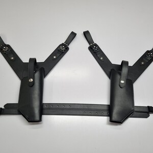 May include: A black dual shoulder holster system featuring two vertical holsters with snap-button retention straps, connected by adjustable shoulder straps and a horizontal waist belt with silver-toned metal buckles and hardware throughout.