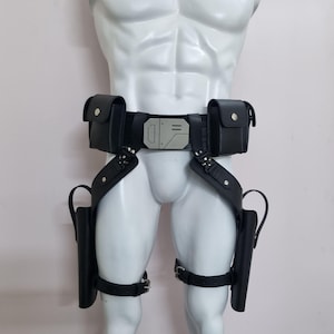 May include: A black utility belt featuring two rectangular pouches, a central metallic buckle with geometric engravings, and two drop-leg holsters attached to the main belt with adjustable straps and silver-toned metal hardware.