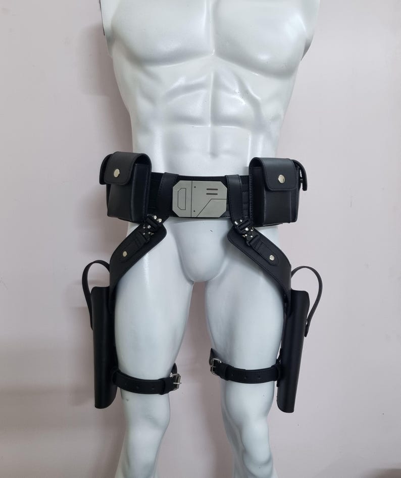 May include: A black utility belt featuring two rectangular pouches, a central metallic buckle with geometric engravings, and two drop-leg holsters attached to the main belt with adjustable straps and silver-toned metal hardware.