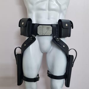 May include: A black utility belt featuring two rectangular pouches with silver snap closures, a central metallic buckle with geometric engravings, and two drop-leg holsters attached to adjustable thigh straps with silver metal buckles.