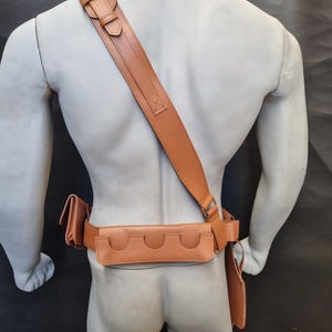 May include: A brown leather belt with a holster and a shoulder strap. The belt has multiple pouches and a buckle.