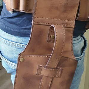 May include: A brown leather holster with a strap and a metal button. The holster is worn on a person's hip.