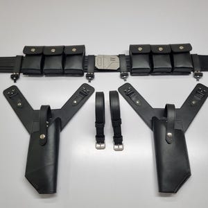 May include: A black utility belt featuring six rectangular pouches with snap closures and a central metallic buckle. Below the belt are two black holster attachments with Y-shaped straps and two separate black adjustable straps with metal buckles.