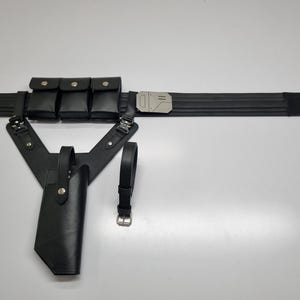 May include: A black utility belt featuring three rectangular pouches with snap closures, a metallic silver buckle, and an attached holster assembly with a secondary strap. The set includes a separate black belt with a silver metal buckle.