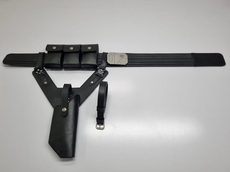 May include: A black utility belt featuring three rectangular pouches with snap closures, a metallic silver buckle, and an attached holster assembly with a secondary strap. The set includes a separate black belt with a silver metal buckle.