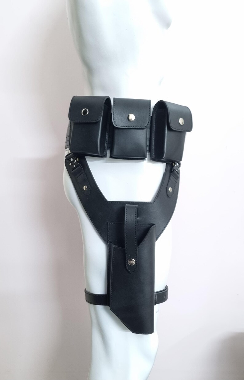 May include: A black leather utility belt featuring three rectangular pouches with silver-tone snap closures attached to the waist, connected to a vertical holster with a strap and snap button, secured by a leg strap around the thigh.