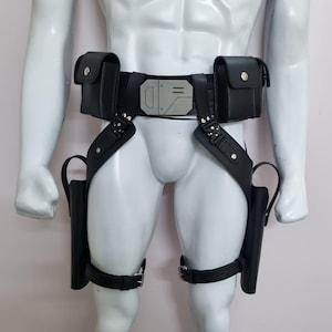 May include: A black utility belt featuring two rectangular pouches, a central silver-toned geometric buckle, and two drop-leg holsters attached with adjustable straps and metal hardware. The design includes a waist belt and thigh straps.