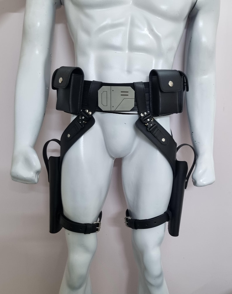 May include: A black utility belt featuring two rectangular pouches, a central silver-toned geometric buckle, and two drop-leg holsters attached with adjustable straps and metal hardware. The design includes a waist belt and thigh straps.