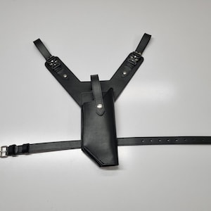 May include: A black leather shoulder holster system featuring a vertical holster pouch, adjustable shoulder straps with metal buckles, and a horizontal waist belt with a silver-toned metal buckle and multiple adjustment holes for a secure fit.