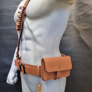 May include: A brown leather belt with a brown leather pouch attached to the right side. The pouch has a flap with a snap closure. The belt has a buckle and a loop for attaching a holster. The belt is worn on a mannequin.