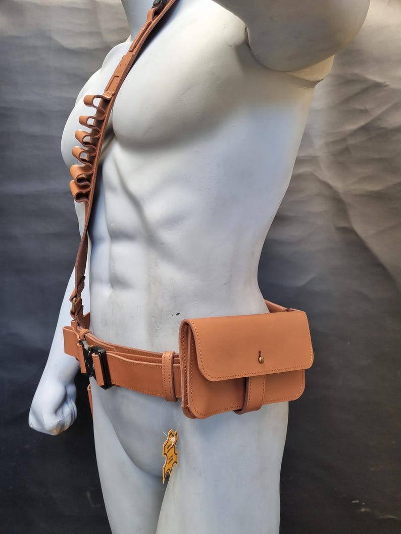 May include: A brown leather belt with a brown leather pouch attached to the right side. The pouch has a flap with a snap closure. The belt has a buckle and a loop for attaching a holster. The belt is worn on a mannequin.