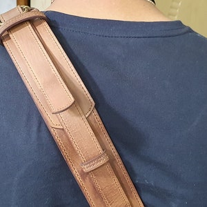 May include: A brown leather shoulder strap with stitching detail. The strap is worn over a person's shoulder.