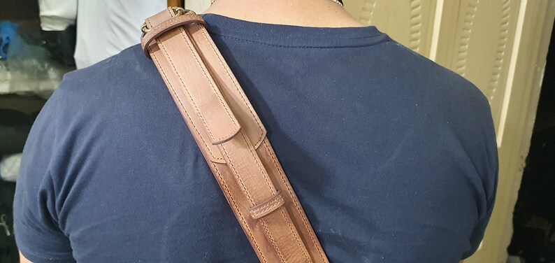 May include: A brown leather shoulder strap with stitching detail. The strap is worn over a person's shoulder.