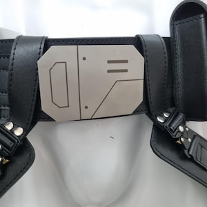 May include: A black utility belt featuring a central metallic buckle with geometric engravings. The belt includes two attached side pouches with snap closures and black tactical-style buckles connecting the lower straps to the main waistband.