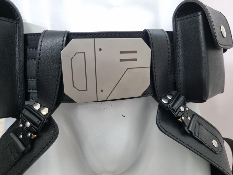 May include: A black utility belt featuring a central metallic buckle with geometric engravings. The belt includes two attached side pouches with snap closures and black tactical-style buckles connecting the lower straps to the main waistband.