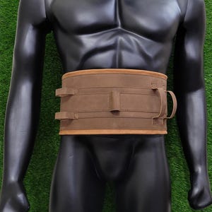 May include: A brown leather belt with two buckles and a strap on the front. The belt is worn on a black mannequin.