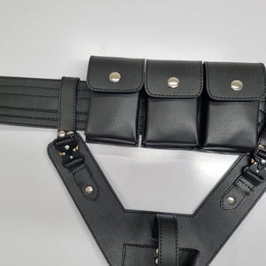 May include: A black utility belt featuring three rectangular pouches with silver-tone snap closures, attached to a structured harness component with black metal buckles and silver rivets. The belt has horizontal stitching and a metallic buckle.
