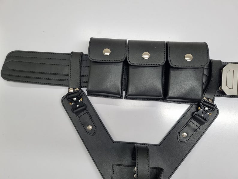 May include: A black utility belt featuring three rectangular pouches with silver-tone snap closures, attached to a structured harness component with black metal buckles and silver rivets. The belt has horizontal stitching and a metallic buckle.