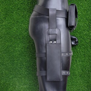 Könnte beinhalten: Schwarzes Leder-Oberschenkelholster mit Gürtel und einer kleinen Tasche. Das Holster hat eine Klappe mit Druckknopfverschluss und ist mit silbernen Nieten verziert.