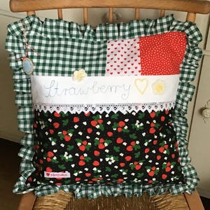 Könnte beinhalten: Ein grünes und weißes Karo-Kissen mit einem weißen Stoffpanel, auf dem "Strawberry" in Kursivschrift mit einer Herz- und Blumenstickerei steht. Das Kissen hat ein schwarzes Stoffpanel mit einem Erdbeer-Druck.