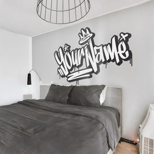 May include: A gray bedroom with a bed and a white wall with a black and white graffiti design that says "Your Name" with a crown above the word "Your".