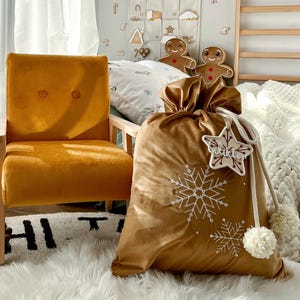 Può includere: Una poltrona in velluto giallo senape con due bottoni sullo schienale, accanto a un grande sacco regalo in tessuto dorato con ricami di fiocchi di neve bianchi, una coulisse bianca con pon-pon e un'etichetta a forma di stella con scritto Wiktor.