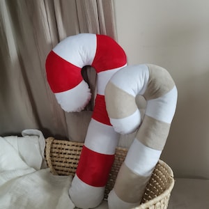 May include: Two plush candy cane pillows, one red and white and one beige and white, are sitting in a woven basket.