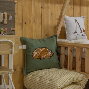 May include: A square olive green throw pillow featuring an embroidered sleeping fawn with white spots. The pillow is placed on a folded yellow and white striped floor mattress with yellow tassels at the corners, near a wooden bed frame.