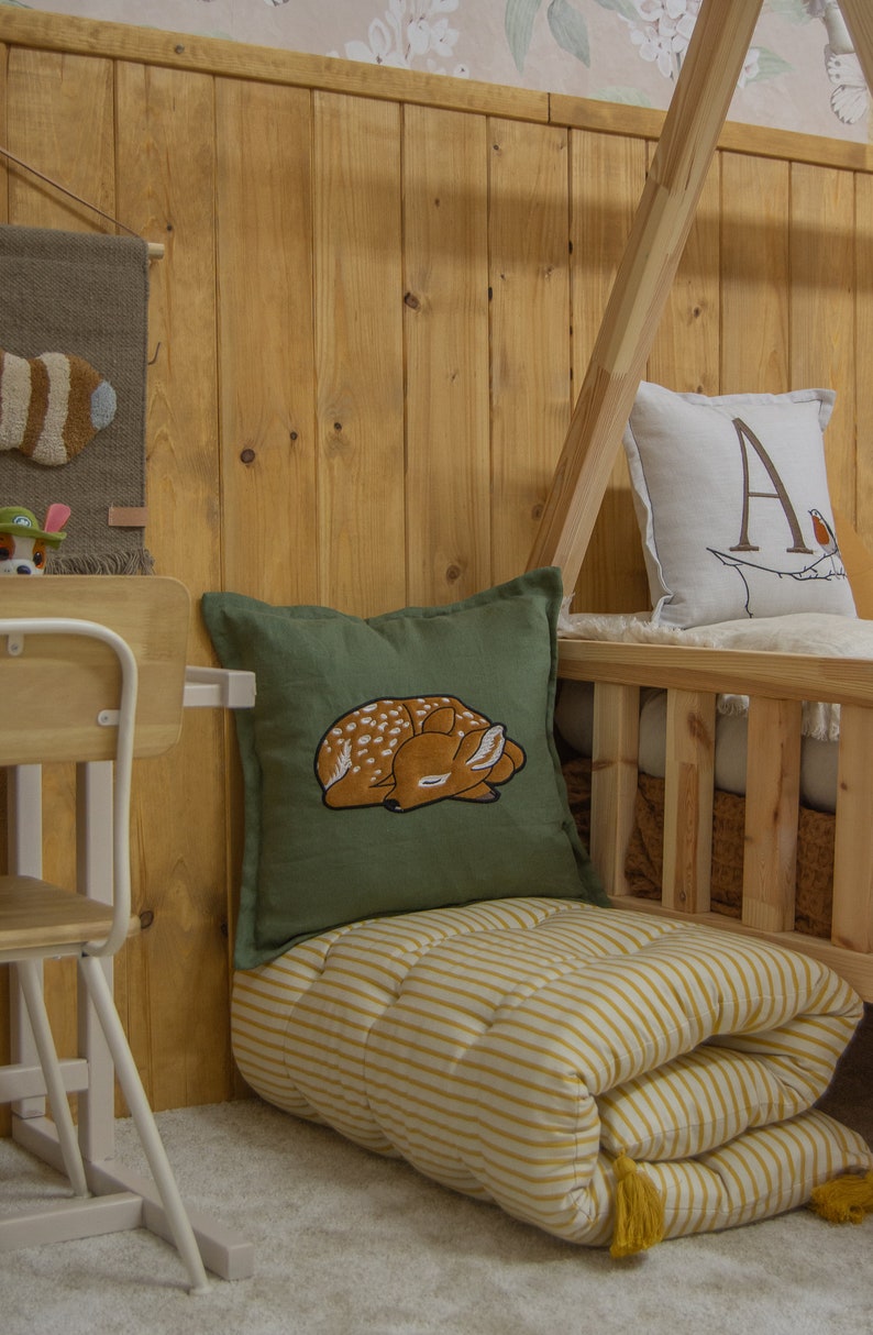 May include: A square olive green throw pillow featuring an embroidered sleeping fawn with white spots. The pillow is placed on a folded yellow and white striped floor mattress with yellow tassels at the corners, near a wooden bed frame.