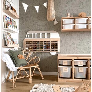 May include: A wall-mounted wooden display shelf shaped like a vintage van with multiple compartments for small toy cars, positioned above a rattan chair with a green pillow, wooden storage units with white bins, and wall-mounted book ledges.