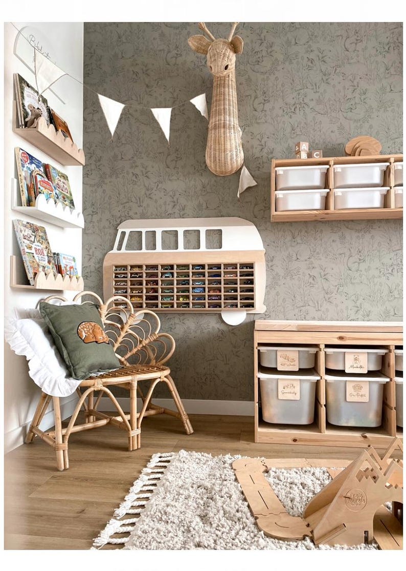 May include: A wall-mounted wooden display shelf shaped like a vintage van with multiple compartments for small toy cars, positioned above a rattan chair with a green pillow, wooden storage units with white bins, and wall-mounted book ledges.