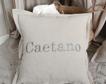 May include: A beige linen pillow with the name "Caetano" embroidered in gray thread. The pillow is lying on a beige fabric with a floral pattern.