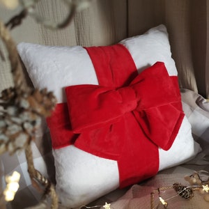 May include: White plush cushion with a large red velvet bow. The cushion is a decorative accent for a home.