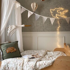 May include: A wooden bed with floral bedding, a green pillow featuring an embroidered fawn, a white canopy, two hanging hot air balloon decorations, a fabric stork mobile, a string of white triangular bunting, and a glowing bird-shaped light.