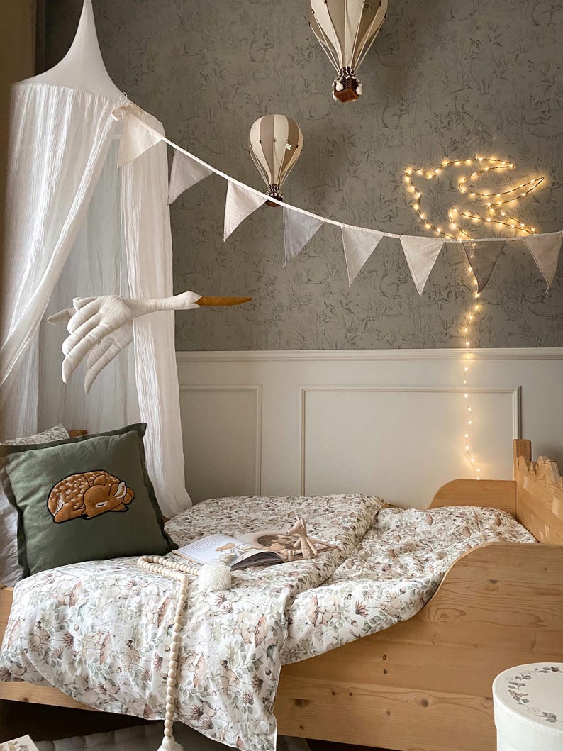 May include: A wooden bed with floral bedding, a green pillow featuring an embroidered fawn, a white canopy, two hanging hot air balloon decorations, a fabric stork mobile, a string of white triangular bunting, and a glowing bird-shaped light.