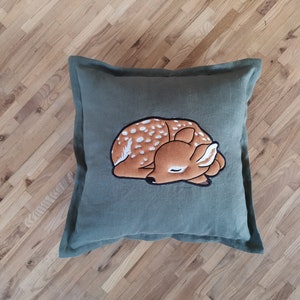 May include: A square olive green throw pillow featuring a central embroidered graphic of a sleeping fawn. The fawn is brown with white spots and a black outline, depicted in a curled, resting position on the solid-colored fabric cover.