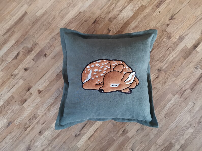 May include: A square olive green throw pillow featuring a central embroidered graphic of a sleeping fawn. The fawn is brown with white spots and a black outline, depicted in a curled, resting position on the solid-colored fabric cover.