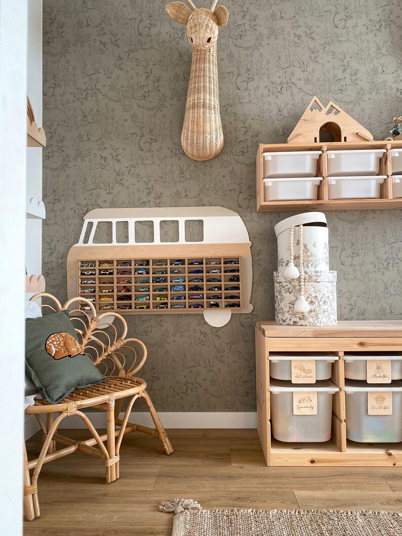 May include: A wall-mounted wooden toy car display case shaped like a vintage van, a rattan chair with a green deer pillow, a wooden storage unit with white bins, a wicker giraffe head wall decor, and stacked round patterned storage boxes.