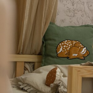 May include: A square olive green throw pillow featuring an embroidered sleeping fawn with white spots. The pillow is placed on a bed with beige bedding, a ruffled edge, and a small brown mushroom-shaped plush toy nearby, with a light beige canopy.