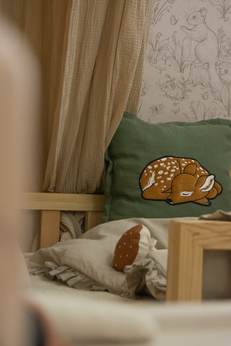 May include: A square olive green throw pillow featuring an embroidered sleeping fawn with white spots. The pillow is placed on a bed with beige bedding, a ruffled edge, and a small brown mushroom-shaped plush toy nearby, with a light beige canopy.
