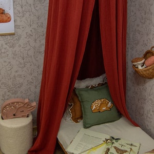 May include: A deep red fabric canopy hangs over a bed featuring a green pillow with a fawn illustration, a white sheet, and an open book displaying various insect drawings. A wooden fawn-shaped lamp sits on a textured white stool nearby.
