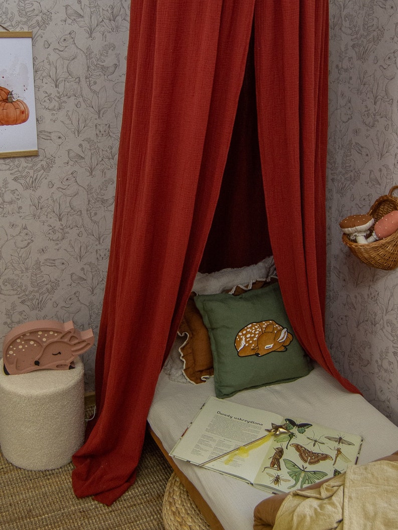 May include: A deep red fabric canopy hangs over a bed featuring a green pillow with a fawn illustration, a white sheet, and an open book displaying various insect drawings. A wooden fawn-shaped lamp sits on a textured white stool nearby.