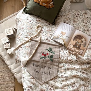 May include: A dark olive green pillow with an embroidered fawn, a beige fabric wall banner with embroidered red berries and the text "Forest is for rest," a wooden bead garland, a storybook with illustrations, and four small wooden blocks.