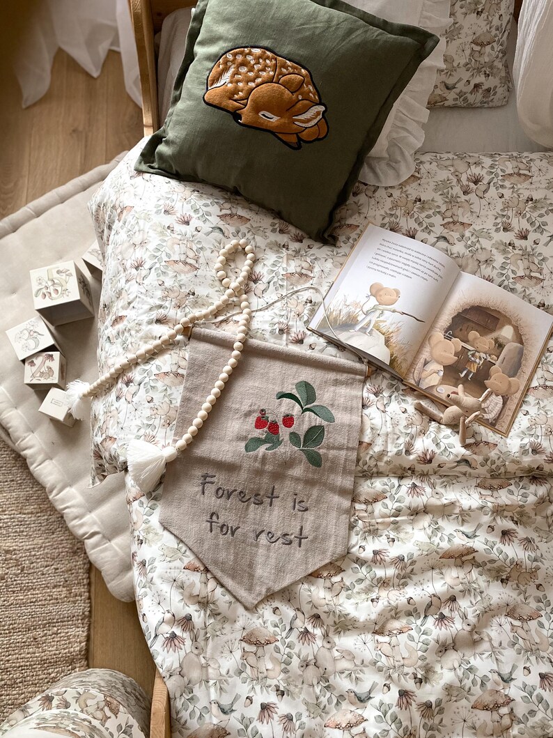 May include: A dark olive green pillow with an embroidered fawn, a beige fabric wall banner with embroidered red berries and the text "Forest is for rest," a wooden bead garland, a storybook with illustrations, and four small wooden blocks.