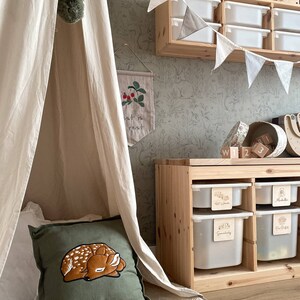 May include: A square olive green throw pillow featuring an embroidered sleeping fawn with white spots. The pillow is positioned inside a cream-colored fabric teepee tent next to a wooden storage unit with white plastic bins and a wall with animal wallpaper.